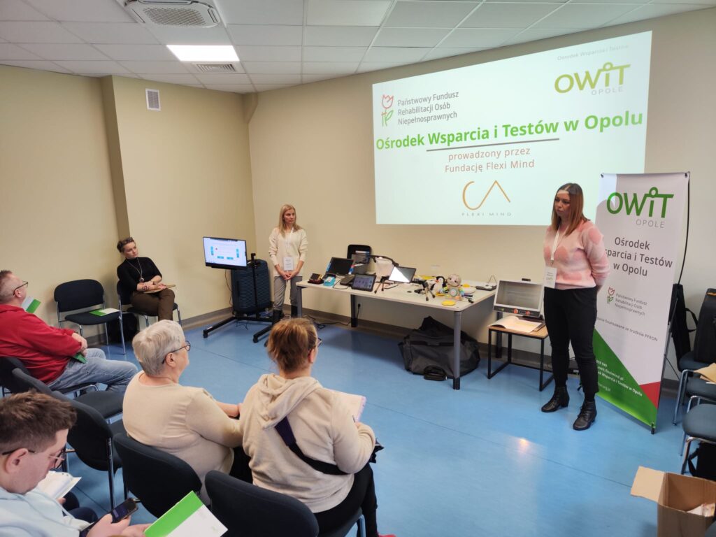 Na środku stoją dwie pracownice OWiT Agnieszka i Kasia. Za nimi na ścianie duży obraz z rzutnika z napisem Ośrodek Wsparcia i Testów w Opolu prowadzony przez Fundację Flexi Mind. Obok napisu jest logo PFRON, OWIT Opole i Fundacji Flexi Mind. Po lewej stronie widocznie z boku i od tyłu osoby siedzące na krzesłach.