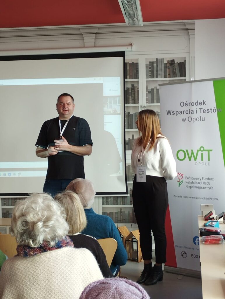 Na środku stoją Rafał i Kasia z OWiT w Opolu. Po prawej stronie baner z logo OWiT Opole i PFRON. Po lewej stronie duży ekran. Widać od tyłu 3 siedzące osoby.