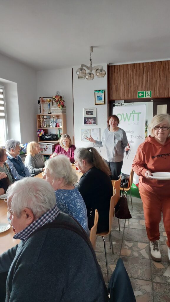 W tle stoi pracownica OWiT w Opolu, za nią baner z logo OWiT w Opolu. Przed nią stoi stół. Po obu stronach siedzą seniorzy i seniorki.