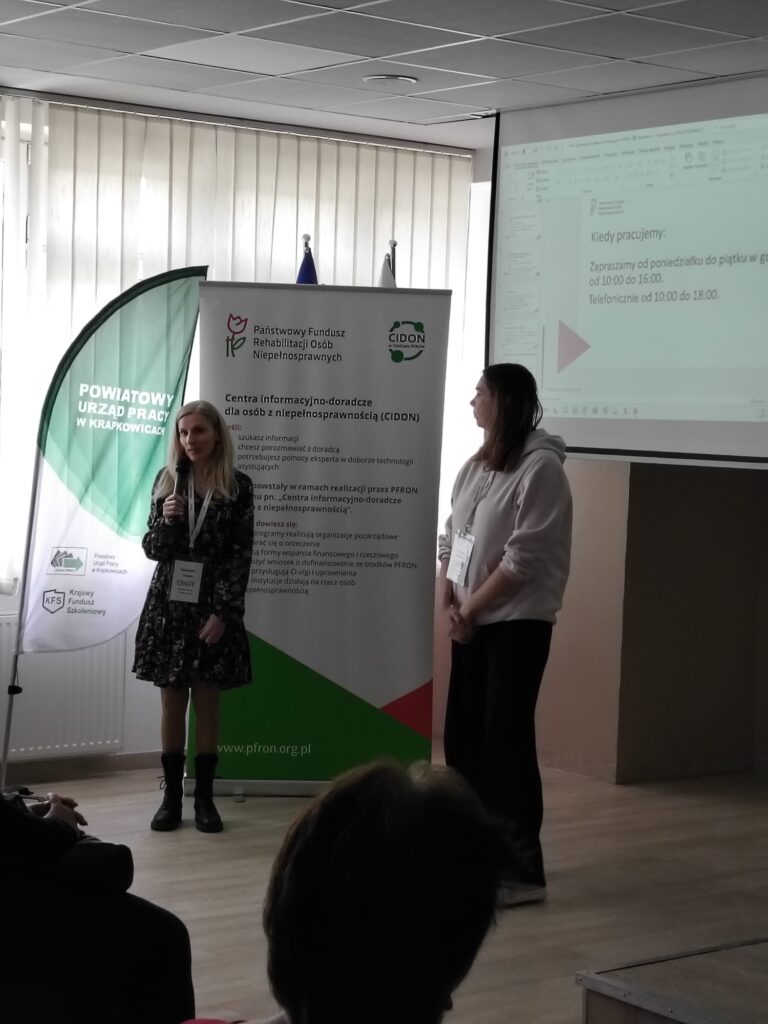 Dwie kobiety stoją przed ekranem, na którym wyświetlana jest prezentacja. Jedna z kobiet mówi do mikrofonu. Za nią banner Centra Informacyjno-Doradcze dla osób z niepełnosprawnością.