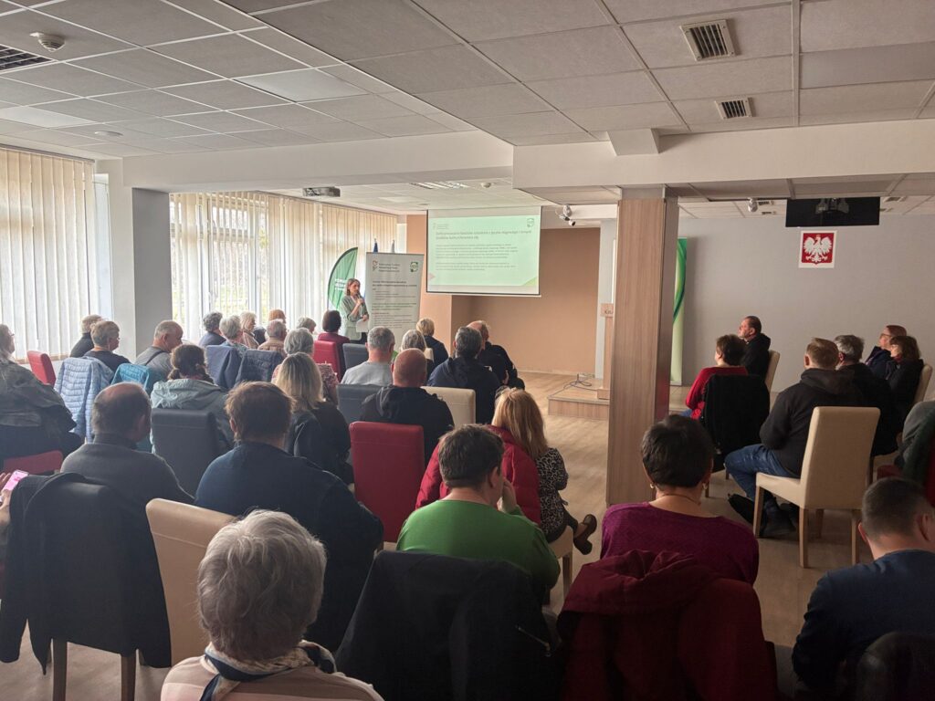 Na pierwszym planie grupa około 30 osób siedzących na krzesłach ustawionych w kilku rzędach. Osoby siedzą tyłem do aparatu. W głębi duży ekran z prezentacją. Po lewej stronie stoi kobieta i mówi do mikrofonu.