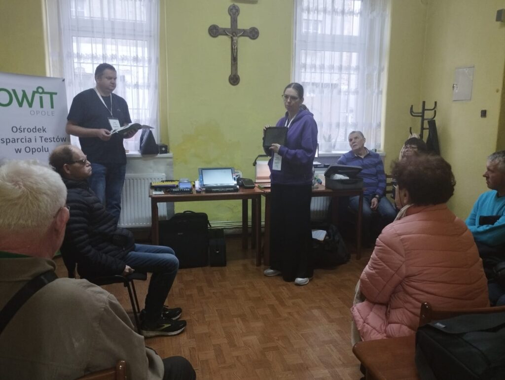 Po prawej stronie stoi kobieta z tabletem w ręce. Po lewej stronie stoi mężczyzna z kartkami w ręce. Za nim stoi baner z logo OWIT Opole. Przed nimi na krzesłach siedzi kilka osób różnej płci, w różnym wieku.