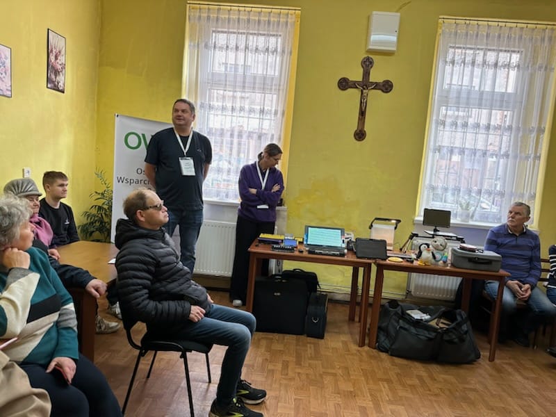 Na stole rozłożone są technologie asystujące. Po prawej stronie stoi siedzi mężczyzna. Po lewej stronie stoi kobieta i mężczyzna. Po lewej stronie pod ścianą siedzi pięć osób na krzesłach.