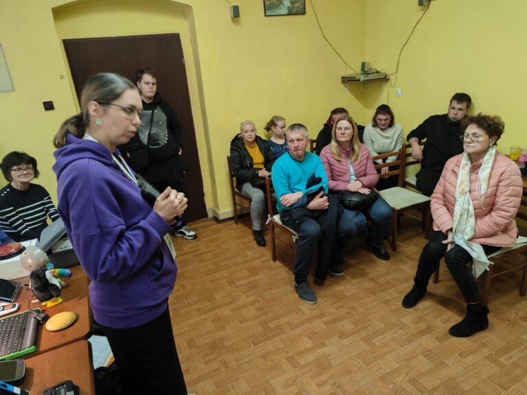 Po lewej stronie stoi kobieta z tabletem w ręce. Za nią na stole stoją technologie asystujące. Po prawej stronie 8 osób w różnym wieku i różnej płci siedzi na krzesłach.