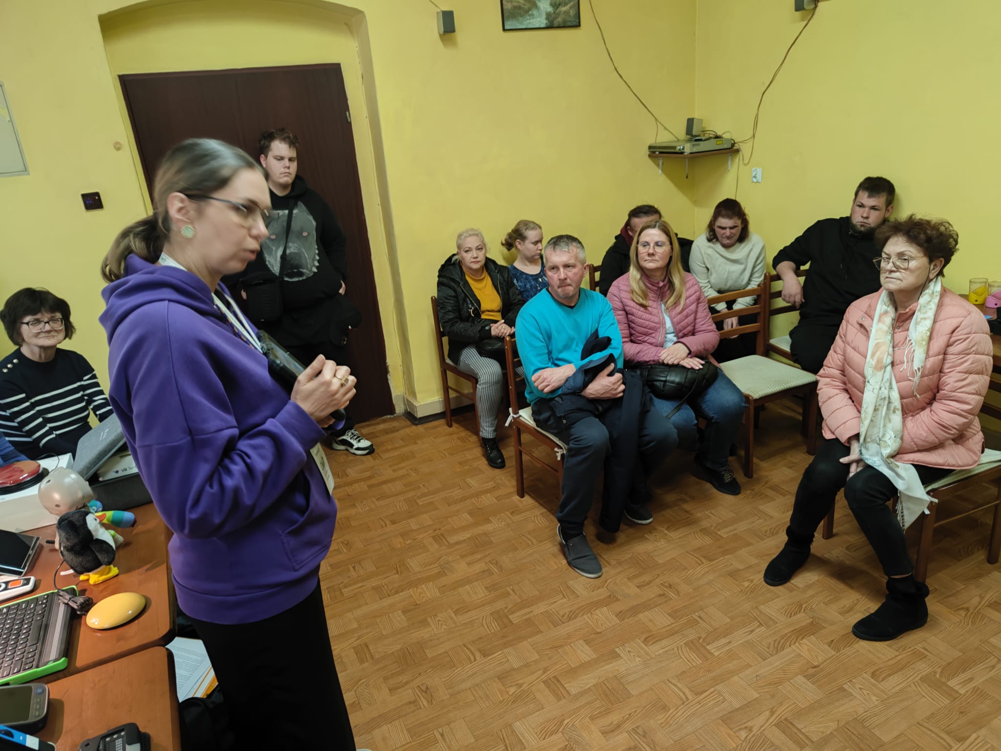 Po lewej stronie stoi kobieta z tabletem w ręce. Za nią na stole stoją technologie asystujące. Po prawej stronie 8 osób w różnym wieku i różnej płci siedzi na krzesłach.