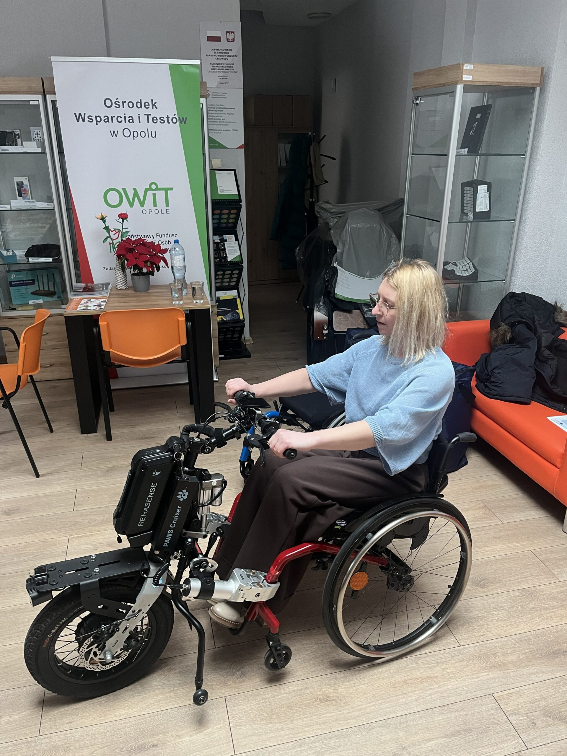 Na wózku siedzi kobieta. Do wózka dołączona jest przystawka elektryczna PAWS Cruiser. Kobieta trzyma przystawkę za rączki.