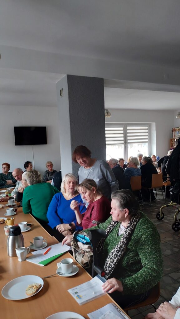 Pracownica OWiT w Opolu rozmawia z seniorkami siedzącymi przy stole. W tle stół w kształcie litery l przy którym po obu stronach siedzą seniorki i seniorzy.