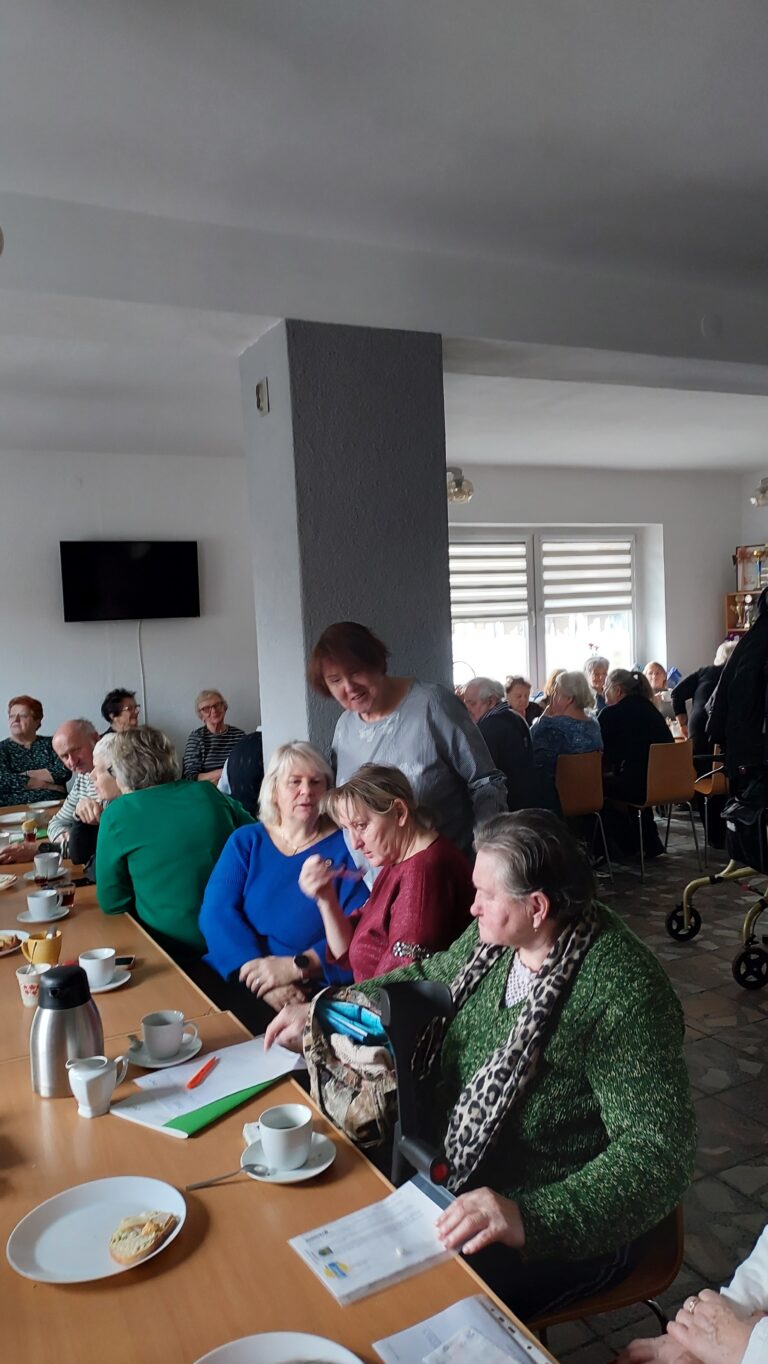 Pracownica OWiT w Opolu rozmawia z seniorkami siedzącymi przy stole. W tle stół w kształcie litery l przy którym po obu stronach siedzą seniorki i seniorzy.