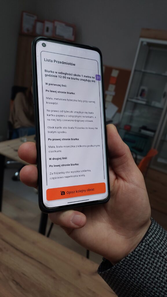 Widoczna prawa dłoń trzymająca smartfon. Na ekranie smartfona widoczny tekst: Lista przedmiotów. Biurko w odległości około metra na godz. 12.00. Na biurku znajdują się: w pierwszej linii: po prawej stronie biurka: mała, metalowa łyżeczka leży przy samej krawędzi. Na prawo od łyżeczki znajduje się biała kartka papieru z odręcznymi notatkami, a na niej leży czerwono-brązowy ołówek.