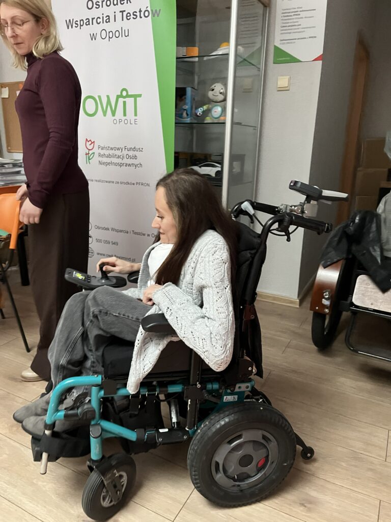 Kobieta siedzi na wózku Action Esprit. Trzyma joystick sterujący wózkiem. Za wózkiem stoi druga kobieta. Po jej prawej stronie banner z logo OWiT Opole.