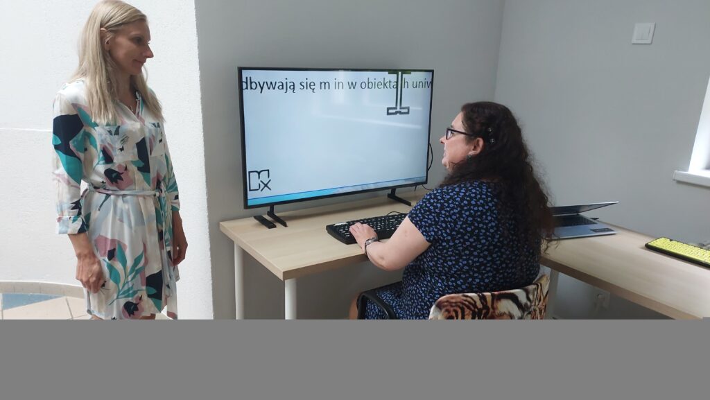 Po lewej stronie stoi pracownica OWiT w jasnej sukience. Po prawej stronie przy biurku siedzi kobieta w niebieskiej sukience. Przed nią leży klawiatura oraz stoi duży ekran. Na ekranie widać słowa napisane dużą czcionką i powiększony kursor