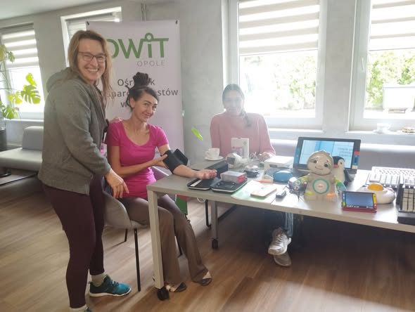 Po prawej stronie jest stół na którym stoją technologie asystujące: system FM, powiększalnik, lupa elektroniczna, powiększona myszka, klawiatury specjalistyczne, zabawka leniwiec. Po lewej stronie stołu siedzi na krześle kobieta i mierzy sobie ciśnienie ciśnieniomierzem głośnomówiącym. Po jej prawej stronie siedzi pracownica OWiT. Po jej lewej stronie stoi kobieta w okularach.