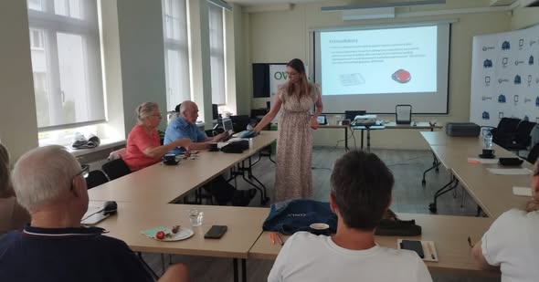 Przy stołach ustawionych w podkowę siedzi 5 osób. Po lewej stronie pracownica OWiT pokazuje urządzenie do komunikacji alternatywnej Smooth Talker