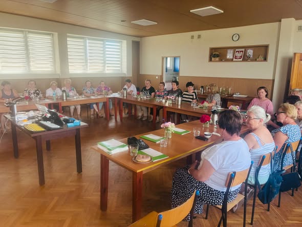 Sala ze stołami ustawionymi w podkowę. Wokół stołów na krzesłach siedzi 18 pań. Na stołach leżą katalogi sprzętu OWiT w Opolu oraz stoją termosy i filiżanki. Po jej lewej stronie stoi stół z technologiami asystującymi jak lupa elektroniczna, klawiatury z kontrastowymi klawiszami, switche, powiększona myszka.