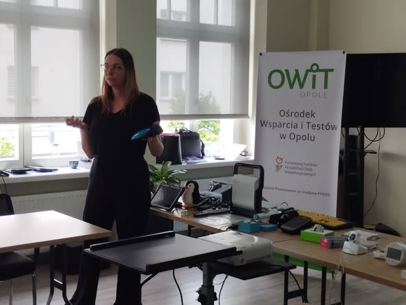 Po lewej stronie stoi pracownica OWiT, trzyma w ręku urządzeni do komunikacji alternatywnej Smooth Talker. Po prawej stronie jest stół na którym są technologie asystujące jak urządzenie lektorskie, mówiący ciśnieniomierz, klawiatura z klawiszami w kontrastowym kolorze, budzik wibrujący i głośnomówiący, tablet, udźwiękowiony zegarek na rękę. W tle rollbanner z napisem Ośrodek Wsparcia i Testów w Opolu