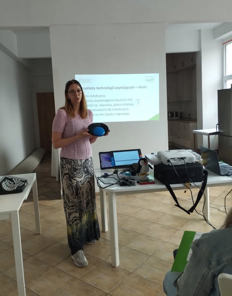 Po lewej stronie stoi pracownica OWiT, trzyma w ręku urządzenie do komunikacji alternatywnej Smooth Talker. Po prawej stronie jest stół na którym są technologie asystujące jak lupa elektroniczna, zabawka Pingwin. W tle ekran z napisem: Przykłady technologii asystujących - słuch.