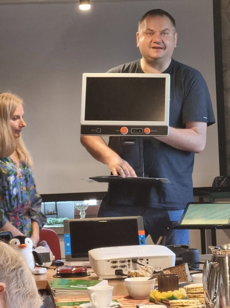 Po prawej stronie stoi pracownik OWIT, trzyma w ręku powiększalnik przenośny. Po lewej stronie stoi pracownica OWiT. Przed nimi stół z lupą elektroniczną i projektorem multimedialnym.