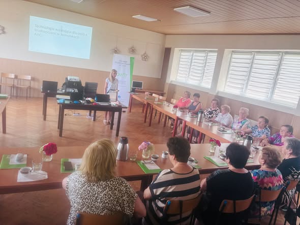 Na pierwszym planie przy stołach ustawionych w kształt litery L siedzi 12 pań. Na stołach leżą katalogi sprzętu OWiT w Opolu oraz stoją termosy i filiżanki. W tle stoi pracownica OWiT. Po jej lewej stronie stoi stół z technologiami asystującymi jak lupa elektroniczna, switche, powiększona myszka. Za nią po prawej stronie stoi rollbanner z logo OWiT w Opolu.