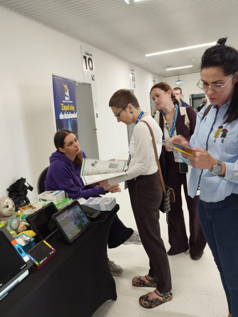 Po lewej stronie stół pokryty czarną tkaniną. Na stole od lewej stoją: myszka powiększona, powiększalnik Traveller, lupa elektroniczna Ruby, tablet z aplikacją Mówik, ciśnieniomierz udźwiękowiony, słuchawki kostne. Na końcu stołu na krześle siedzi pracownica OWiT. Przed nią stoi osoba, przegląda katalog technologii asystujących. Obok niej stoi kobieta i patrzy do katalogu. Po prawej stronie stoi kobieta i czyta ulotkę