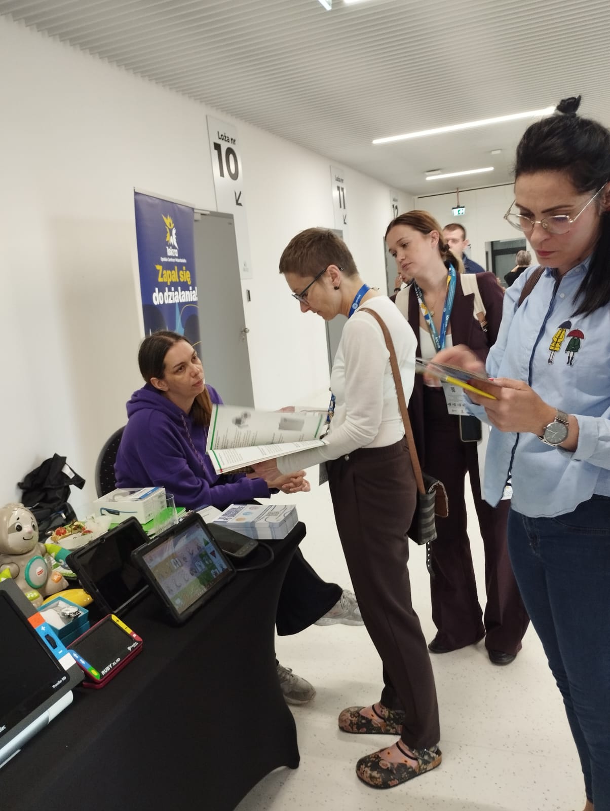 Po lewej stronie stół pokryty czarną tkaniną. Na stole od lewej stoją: myszka powiększona, powiększalnik Traveller, lupa elektroniczna Ruby, tablet z aplikacją Mówik, ciśnieniomierz udźwiękowiony, słuchawki kostne. Na końcu stołu na krześle siedzi pracownica OWiT. Przed nią stoi osoba, przegląda katalog technologii asystujących. Obok niej stoi kobieta i patrzy do katalogu. Po prawej stronie stoi kobieta i czyta ulotkę