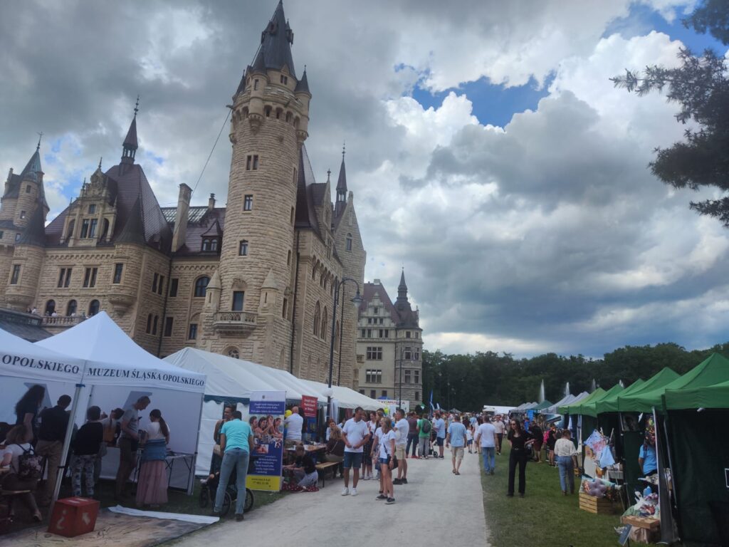 Po lewej stronie widoczne mury zamku w Mosznej. Na wprost szeroka aleja. Po obu stronach alei stoją namioty ustawione obok siebie. Przed namiotami i w namiotach tłum osób.