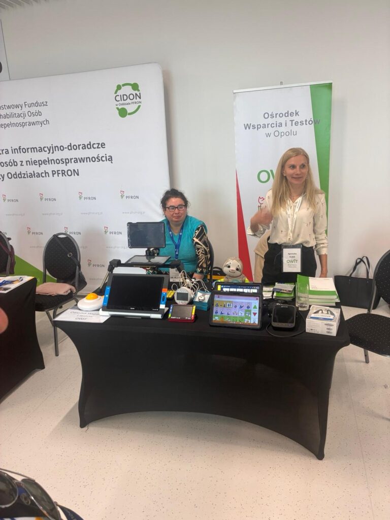 Stół pokryty czarną tkaniną. Na stole od lewej stoją: myszka powiększona, powiększalnik Traveller, lupa elektroniczna Ruby, tablet z aplikacją Mówik, ciśnieniomierz udźwiękowiony, słuchawki kostne. Za stołem po prawej stronie stoi pracownica OWiT, po lewej stronie siedzi stażystka OWiT. Za nimi stoi rollup z napisem Ośrodek Wsparcia i Testów w Opolu