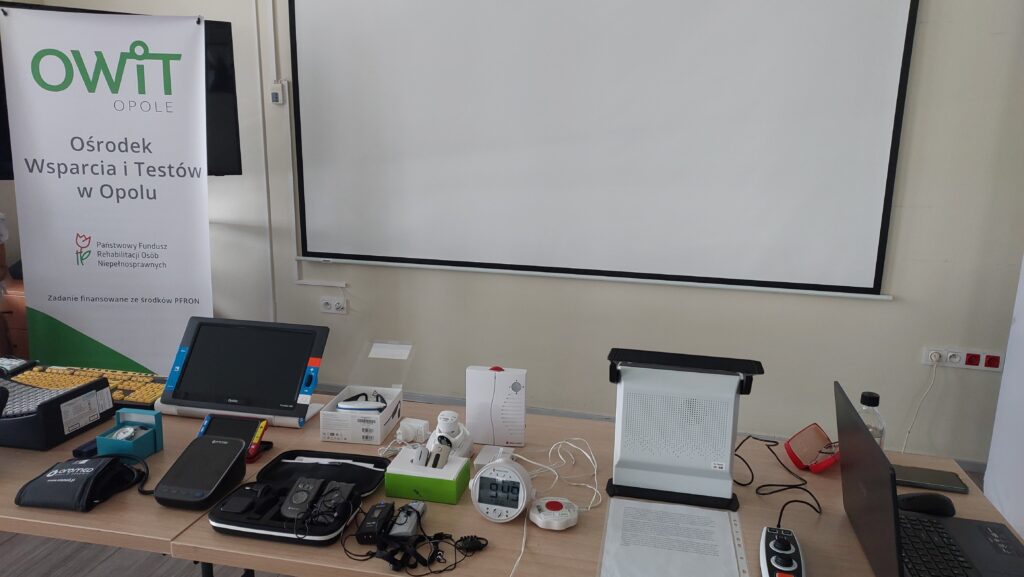 Stół na którym są technologie asystujące jak urządzenie lektorskie, mówiący ciśnieniomierz, klawiatura z klawiszami w kontrastowym kolorze, budzik wibrujący i głośnomówiący, lupu elektroniczne, system FM, udźwiękowiony zegarek na rękę. W tle po lewej stronie rollbanner z napisem Ośrodek Wsparcia i Testów w Opolu