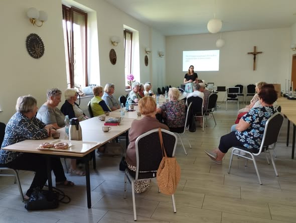 Na pierwszym planie po lewej stronie stoi długi stół. Wokół niego na krzesłach siedzi ponad 16 osób. Siedzą tyłem do obiektywu. W tle stoi pracownica OWiT.