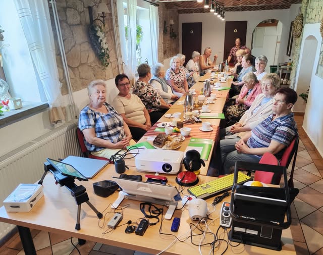 Na pierwszym planie jest stół na którym są technologie asystujące jak urządzenie lektorskie, mówiący ciśnieniomierz, klawiatura z klawiszami w kontrastowym kolorze, budzik wibrujący i głośnomówiący, tablet, powiększalnik Exigo, słuchawki kostne, system FM. Za nim wokół stołów ustawionych w jednym rzędzie siedzi 18 kobiet, w tle stoi jedna kobieta. Na stołach stoją termosy, filiżanki, talerze z ciastem.